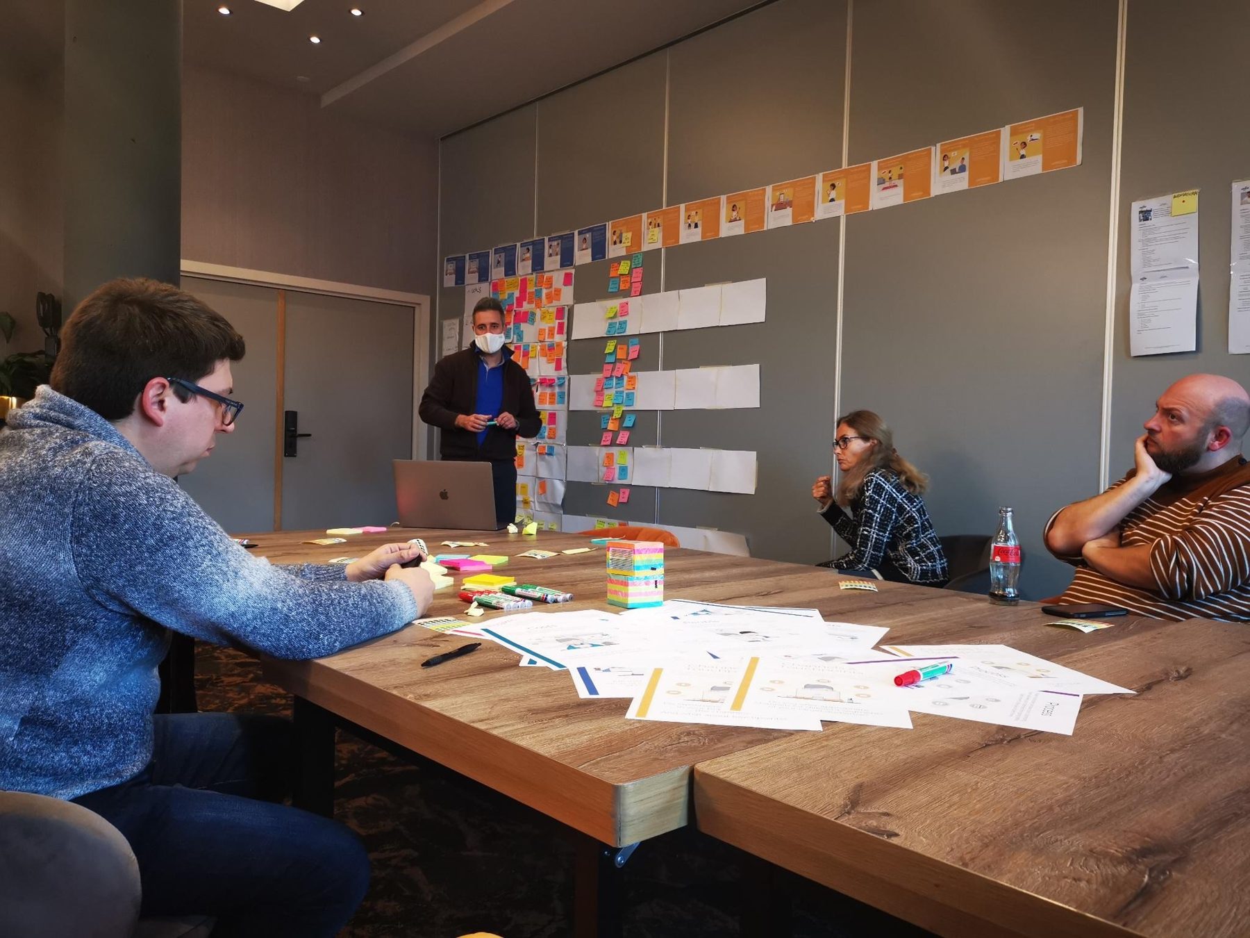 L'atelier sur place : l'animation devant le mur de personas et de post-its, les participants autour de la table.